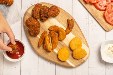 Fried potatos and nuggets served on a wooden board on a white wooden table. More ingredients surround them: ketchup and mayonnaise, sliced tomatoes and chilli pepper. A hand is dipping a nugget in ket