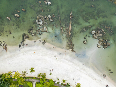 Aerial view of pristine white sands meeting the turquoise sea, scattered with weathered rocks and lush palm trees lining the coast, Hua Hin, Prachuap Khiri Khan, Thailand.