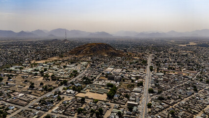 Aerial view of a sprawling cityscape nestled beneath a hazy sky, with a prominent rocky outcrop and telecommunication mast piercing the skyline, Jalingo, Taraba, Nigeria.