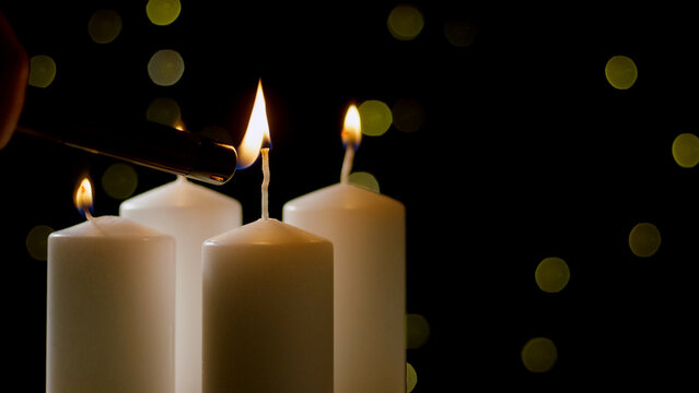 A hand uses a lighter to ignite the wick of a white candle, creating a warm glow against a dark background.