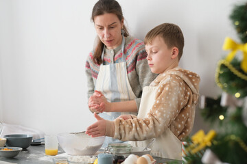 A woman guides her son in baking cookies, mixing flour in a bowl on a kitchen counter decorated for...