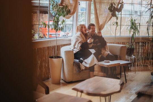 A joyful couple shares drinks and laughter while wrapped in blankets on a comfy sofa.