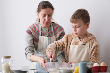 A woman guides her son in baking cookies, mixing flour in a bowl on a kitchen counter decorated for...