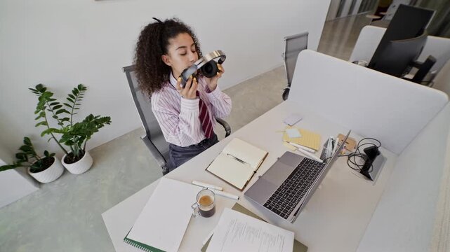 High angle of young female manager in smart casualwear siting by workplace, taking notes in notepad, then taking off headphones, folding laptop, getting up from chair and leaving office