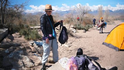 A pretty woman stands on the beach with a backpack, wearing a hat and glasses. Camping by the sea. A group of friends