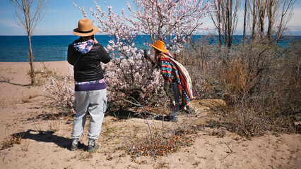 Two women in stylish authentic clothes are standing by a flowering tree by the sea. Two tourists...