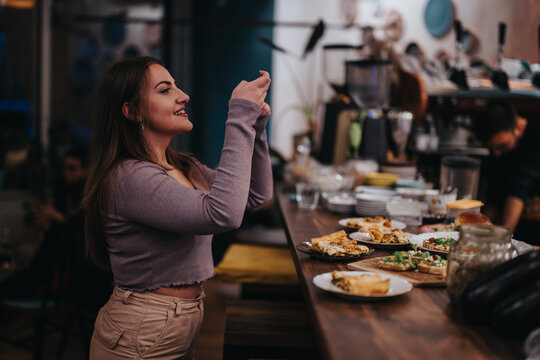 Young woman captures a photo of delicious food served on a wooden counter in a cozy restaurant. The scene highlights culinary presentation, social interaction, and passion for documenting experiences.