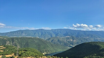 Naklejka premium Breathtaking panoramic shot captures lush green mountains cascading down to a serene lake under a clear blue sky. Ideal for travel, nature, and outdoor themes.