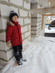 Young boy in vibrant red winter jacket and knit hat leaning playfully against snowy white block...
