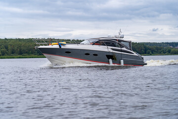 Luxurious grey and white yacht with prominent red stripe cruises dynamically on calm river under cloudy sky, leaving foamy white wake behind it with lush green treeline in background.
