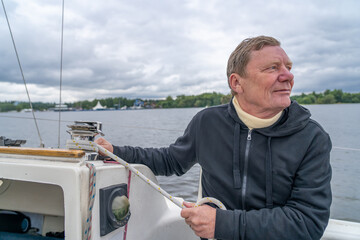 Active senior man skillfully managing sailboat's ropes on winch, looking thoughtfully across calm...
