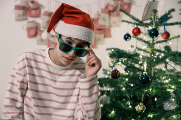 Cool teenager girl with Santa hat and sunglasses posing near decorated Christmas tree during winter holidays