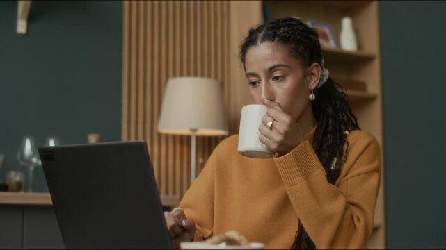 Medium shot of concentrated young biracial girl sitting at table in kitchen and working on laptop sipping coffee or tea from mug