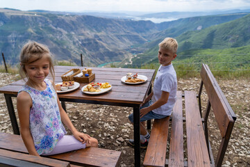 Young siblings enjoying delightful outdoor breakfast at picnic table, set against breathtaking panoramic backdrop of majestic mountains, deep valleys, and winding turquoise river below.