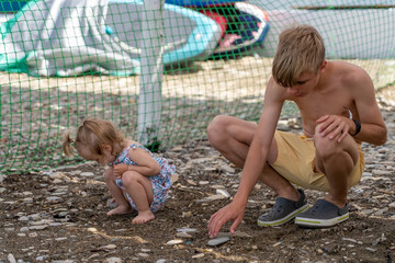 Sibling bond: young boy and his little sister engaging in quiet moment, crouching on textured ground, observing stones by beach