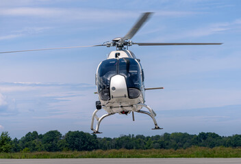 A white Airbus H120 helicopter captured head-on while hovering above the runway. Rotor blades blur with motion against a layered sky, while green trees and open grassland frame the horizon below. © Paul