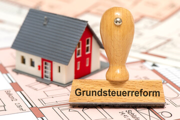 Ein Holzstempel ist beschriftet mit Grundsteuerreform über einem Bauplan mit Modellhaus als...