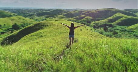 Young woman standing on a lush green hill with open arms, embracing the stunning landscape of Sumba Island in Indonesia, feeling the warmth of the sun and the gentle breeze