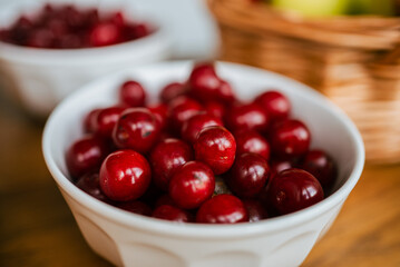 Ripe red cherries overflowing a white ceramic bowl on a rustic wooden table, with another bowl and a basket of fruit softly blurred in the background