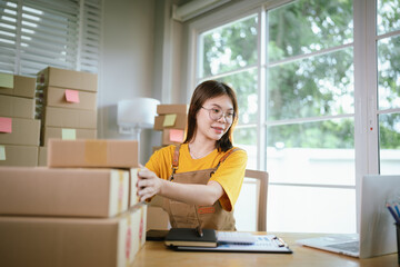 A young Asian female entrepreneur works from home, managing online sales, shipping parcels, marketing products, and ensuring client satisfaction efficiently.