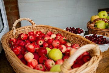 Wicker basket overflowing with small red and yellow crab apples rests on a wooden surface, accompanied by bowls of cherries and a basket of larger apples, reflecting autumn's bounty