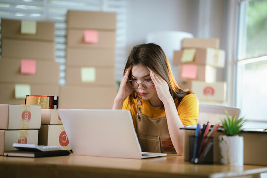 A young Asian businesswoman sits stressed at her desk, managing online orders, shipping boxes, facing retail challenges, worry, and frustration.