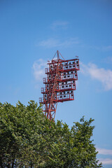Bright floodlights stand tall against blue sky and greenery during early afternoon at a sports...