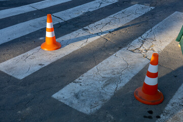Two orange traffic cones placed on a damaged pedestrian crossing with visible cracks and shadows in...