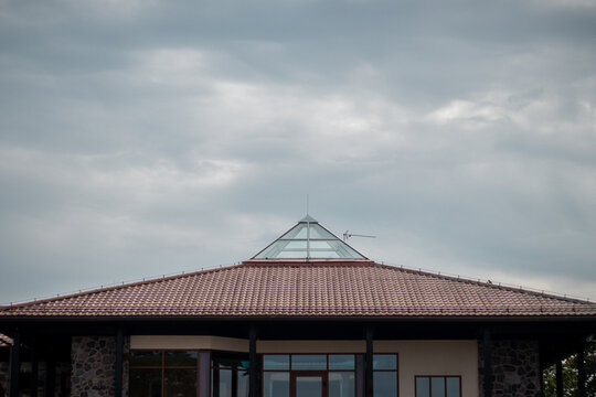 A contemporary house featuring a red-tiled roof and a central glass pyramid skylight under a cloudy, overcast sky. - Powered by Adobe