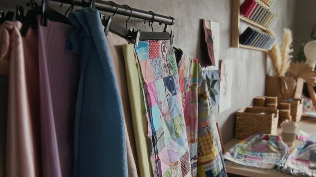 Medium shot of rail with various handmade merchandise on wall at sewing workshop or manufactory, with silk carves, patchwork jacket, part of unfinished quilt, colorful bobbins of thread on wall