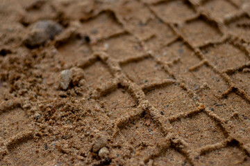 Detailed macro view of a vehicle tire tread imprint in sandy dirt, showing texture and geometric patterns left on the ground.