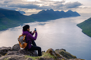 A traveler sits on rocky terrain in the Faroe Islands, taking a picture of the stunning landscape....