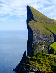 A solitary figure stands on a lush green Mylingur cliff, overlooking a striking rock formation that...