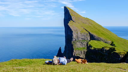An Asian woman basks in the sunlight on a grassy Mylingur cliff in the Faroe Islands, enjoying breathtaking ocean views and the majestic rock formations that rise sharply from the sea.