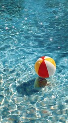 Obraz premium Colorful red, yellow, and white beach ball creates gentle ripples as it floats in the inviting, shimmering blue water of a resort swimming pool on a sunny day.