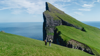 Two hikers stand on the lush green hills overlooking the dramatic cliffs of the Faroe Islands. The...