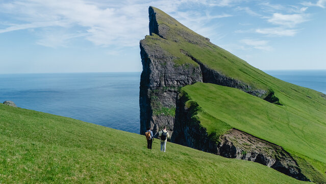 Two hikers stand on the lush green hills overlooking the dramatic cliffs of the Faroe Islands. The vast ocean stretches beyond, under a clear blue sky, creating a tranquil and inspiring atmosphere.