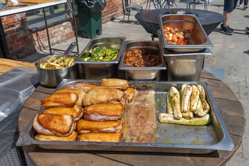 Assortment of grilled meat, zucchini, and cooked vegetables in stainless steel trays on outdoor wooden table.