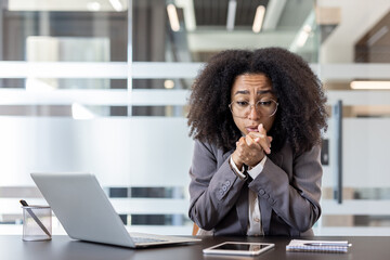 Thoughtful and upset young African American woman in a suit sitting at a table with a laptop in the...