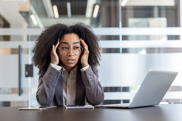 Tired and overwhelmed young African American woman sitting at her desk in the office and holding...