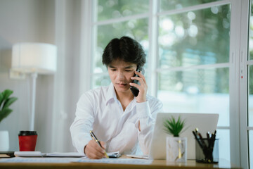 Obraz premium An Asian businessman in a white shirt works at his office desk, analyzing charts and reports with laptop, clipboard, and documents.