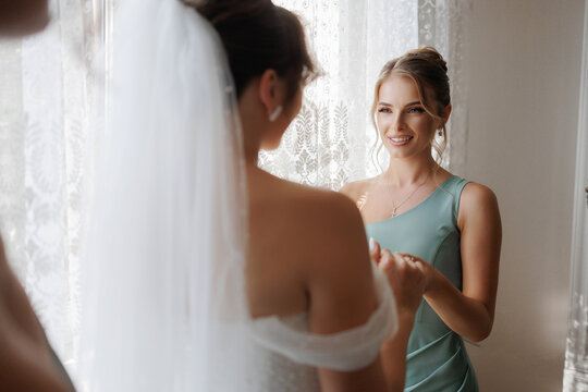 Bridesmaid helps bride prepare for wedding in a bright, elegant room