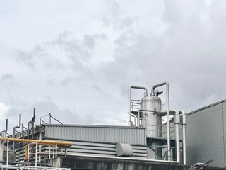 Obraz premium the top of an industrial building featuring a large, cylindrical tank connected to a network of pipes. The machinery is part of a factory or plant, set against a cloudy sky, highlighting the intriguin