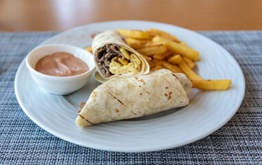 Delicious burrito with beef and eggs served with crispy fries and dipping sauce on a light-colored plate