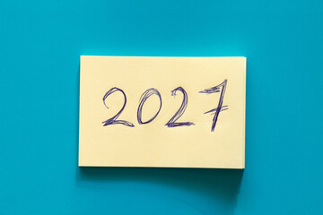Yellow sticky notepaper with large handwritten 2027 inscription in blue ink on blue background on the theme of new year and plans, date