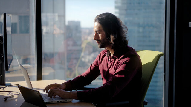 Millennial caucasian man working on a laptop by a large window in a bright modern office, focused and productive, highlighting technology, communication and professional dedication indoors. - Powered by Adobe