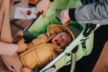 A scene depicting a young child crying while two adult hands prepare them in a stroller. The...
