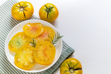 Fresh yellow tomatoes sliced on a white plate with rosemary garnish, surrounded by whole tomatoes,...