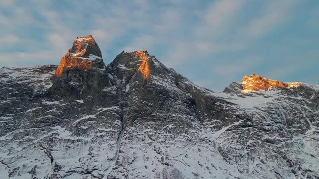 This drone footage captures the iconic snowy peaks of Trollstigen in Norway, bathed in the warm, golden light of sunrise or sunset.