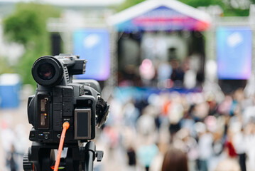 large black video camera on tripod outdoors, shooting music festival and crowd of people outdoors,...